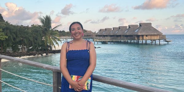 Woman in blue dress smiling by tropical waterfront with overwater bungalows.