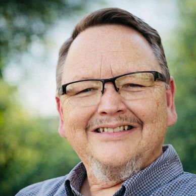 Smiling man wearing glasses outdoors with a blurred green background.