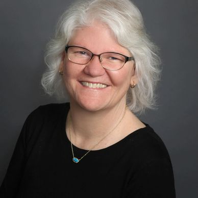 Smiling woman with white hair and glasses against a gray backdrop.