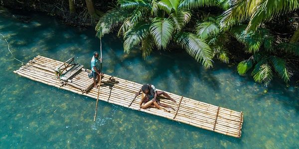 Two people on a bamboo raft floating on clear blue water near lush green foliage.