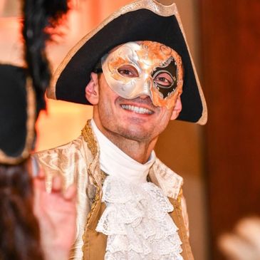 Man in an ornate masquerade mask and 18th-century costume smiling.