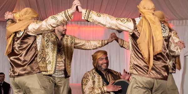 Men in traditional attire dancing and playing a drum at a cultural event.
