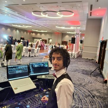 A DJ in a vest and bow tie at a formal event with laptops and DJ equipment.
