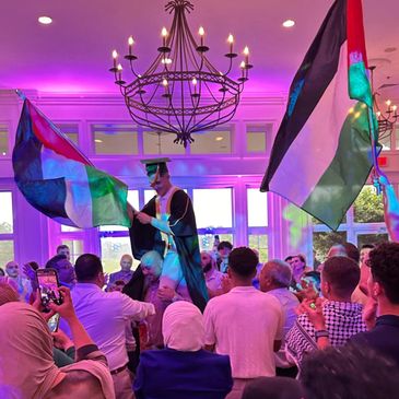 Graduate celebrating with colorful flags and crowd indoors under purple lighting.