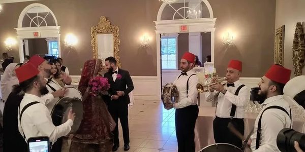 A couple dances surrounded by musicians in traditional hats inside an elegant hall.