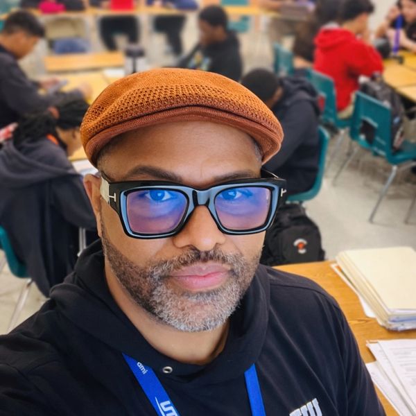 Man wearing a brown cap and large black glasses in a classroom.