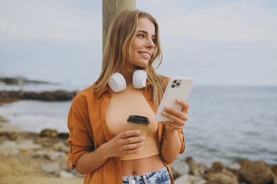 A woman on a beach with a phone and coffee, showing confidence and growth from self-esteem therapy.