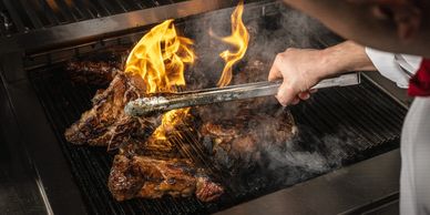 Chef grilling steaks with flames rising on a large grill highlighting dining options.