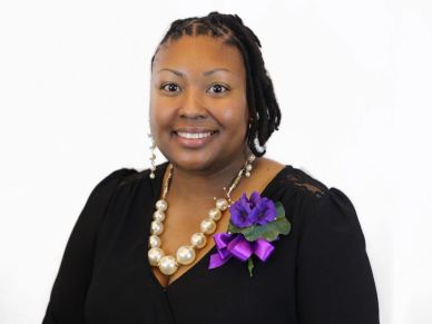 Smiling woman wearing a black dress with pearl necklace and purple flower corsage.