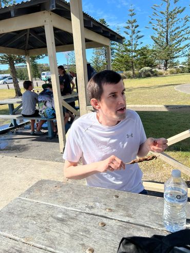 Man in white shirt eating food outdoors at picnic table.