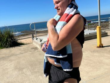 Man with a towel over his shoulder walking near a beach on a sunny day.