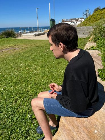 A man in casual clothes sitting on a rock near the ocean under clear skies.