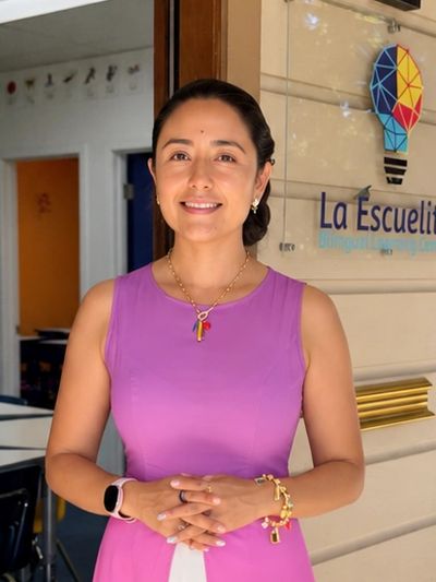 Mujer sonriente vestida de rosa frente a un salón de clases.