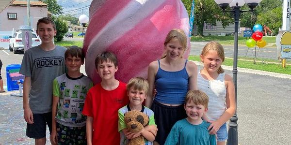 Seven smiling children posing outdoors in front of a large pink sculpture.