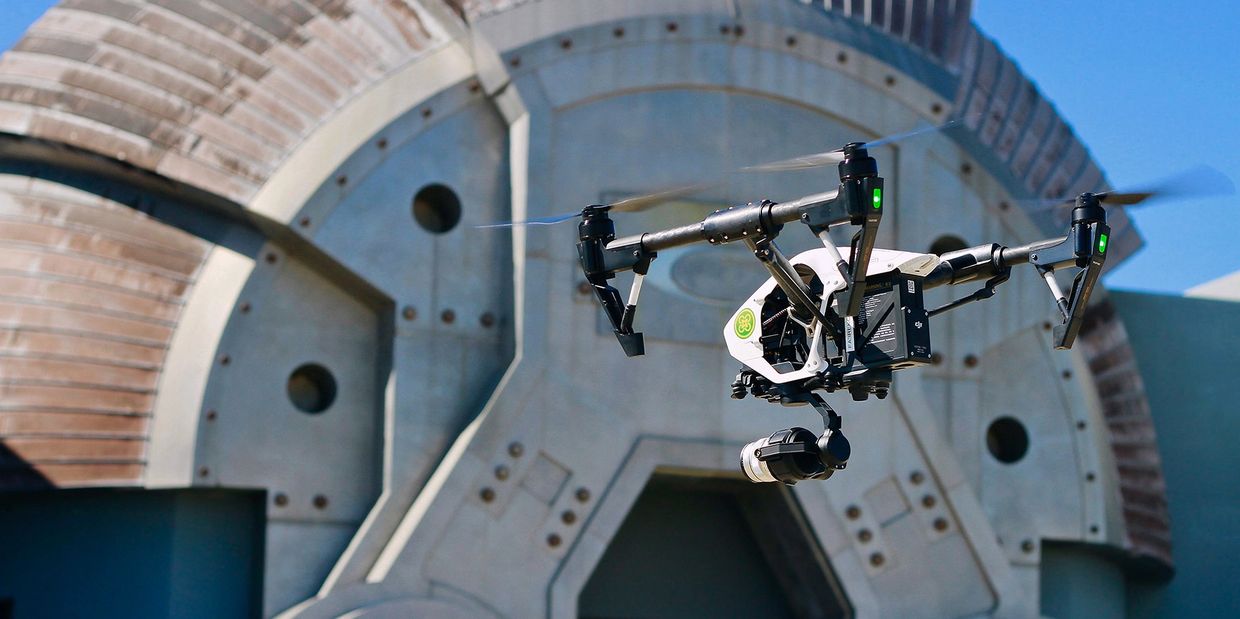 A drone flying in front of a large, industrial-style circular structure under clear blue sky.