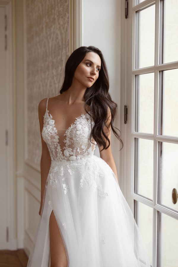 Woman in elegant white lace wedding dress standing by window.