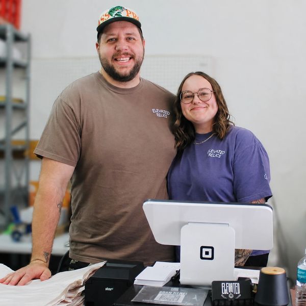 Two smiling people behind a counter with Elevated Relics shirts.