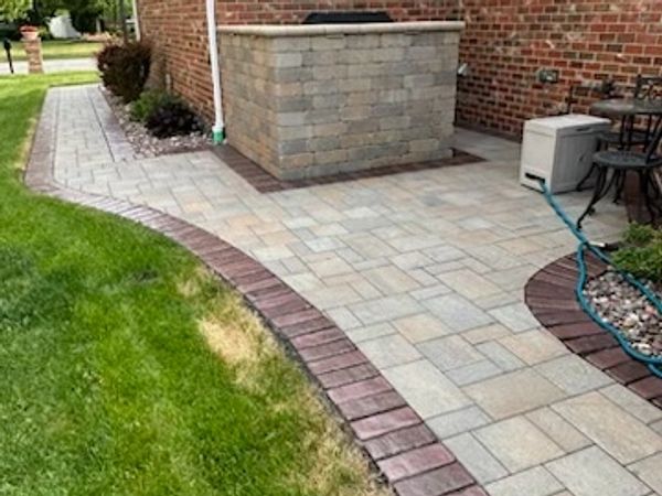 A paved patio with brick edging beside a brick house and green lawn.