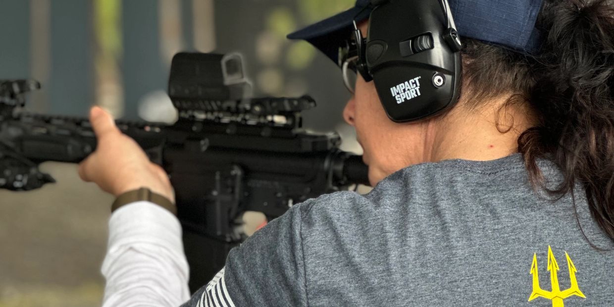Person aiming a rifle while wearing ear protection.