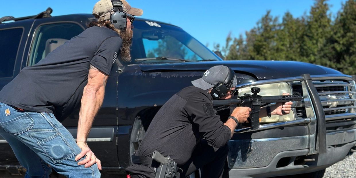Two men at a shooting range using a black pickup for cover.