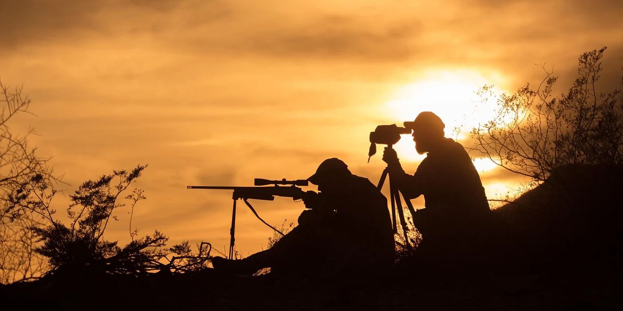Silhouette of two people with a rifle and camera tripod at sunset.
