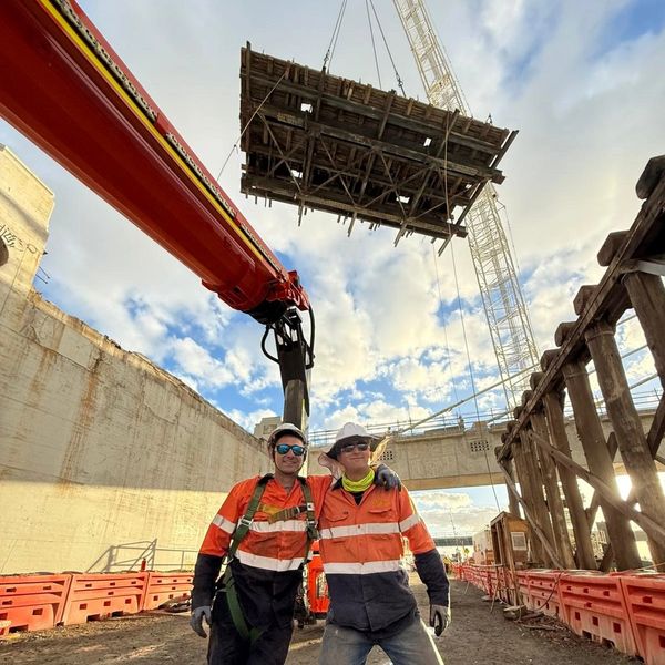 Two construction workers posing inside a construction site with a crane lifting materials.