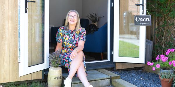 Smiling woman in floral dress sitting at an open door with an "Open" sign and a dog inside.