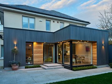 Modern two-story house with wood and metal exterior, large glass windows, and outdoor lighting.