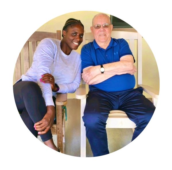 A smiling young woman and an elderly man sitting closely on wooden chairs.
