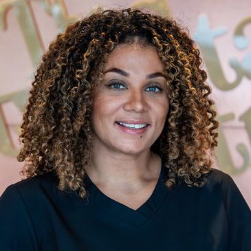 Smiling woman with curly hair in black scrubs at a reception desk.