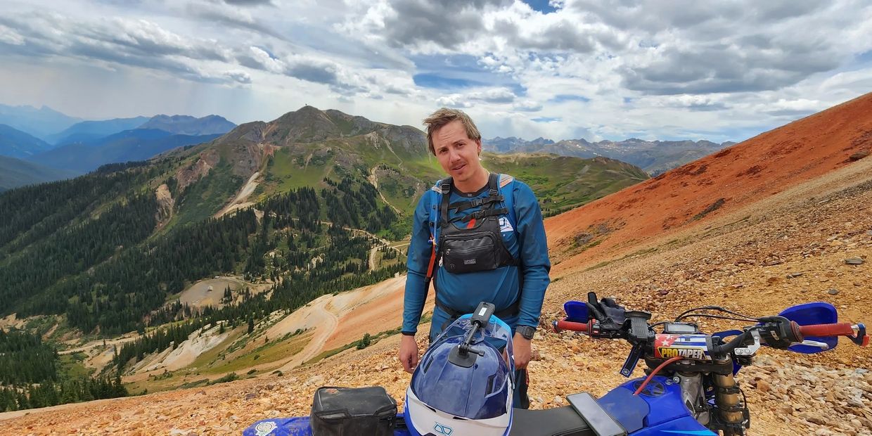 Man stands beside a blue dirt bike on a mountainous trail under a cloudy sky.