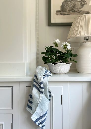 A cozy kitchen corner with a striped cloth, white flowers in a textured pot, and a ceramic lamp.