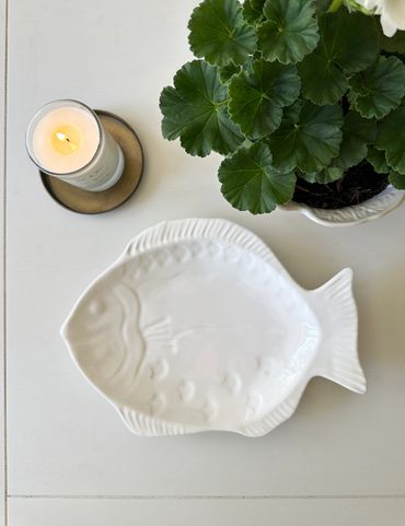 White fish-shaped ceramic dish next to a lit candle and green plant.