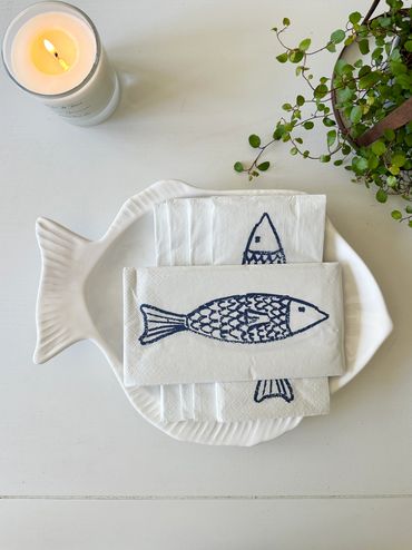 Fish-shaped plate with fish-printed napkins, a candle, and a plant on a white surface.