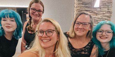 Five smiling women, two with teal hair, posing indoors.