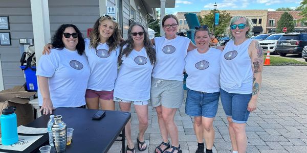 Group of six women in matching white shirts posing outdoors on a sunny day.