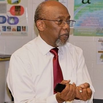 Man in white shirt and red tie speaking and gesturing indoors.