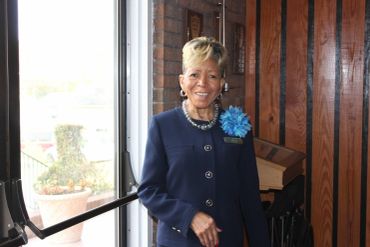 Elegant woman in a navy suit with a blue flower brooch smiles indoors.