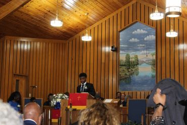 A man speaks at a wooden-paneled church with a scenic mural behind him.