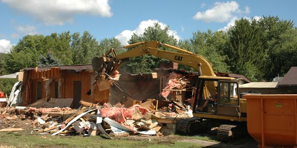 Excavator demolishing a house in a residential area on a sunny day.