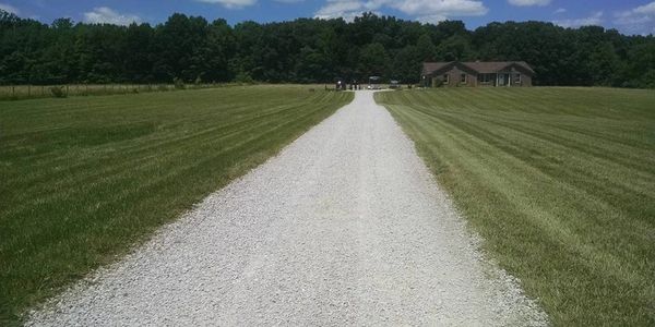 Long gravel driveway leading to a house surrounded by green grass and trees.