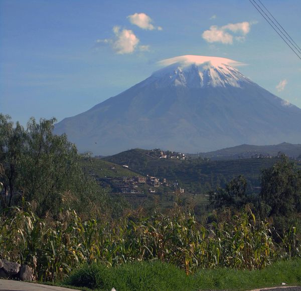 Snow-capped mountain with a lenticular cloud and green fields below.