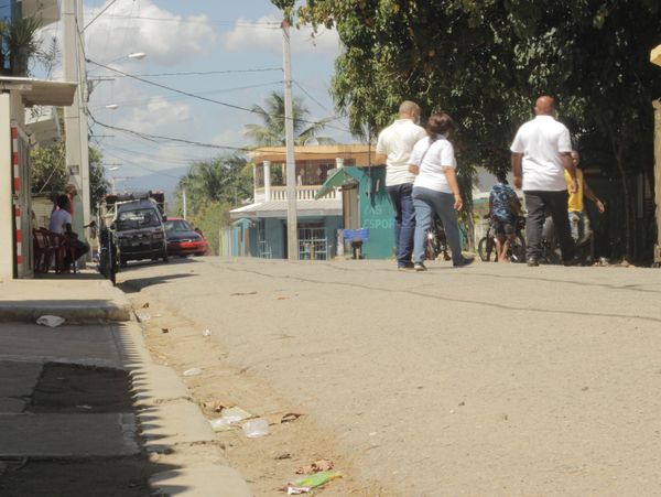 People walking on a sunny street with parked cars and trees.