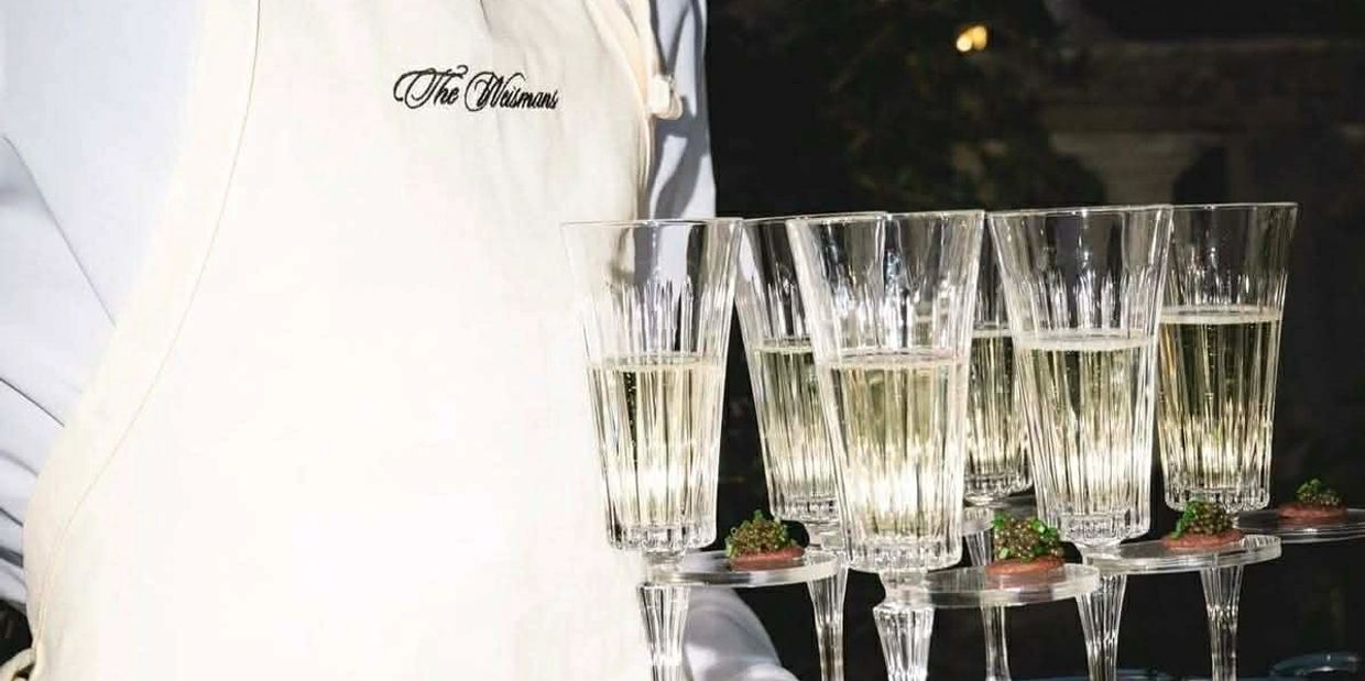 A waiter wearing a "The Weismans" apron carries a tray of champagne flutes with appetizers.