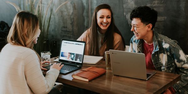 3 pessoas sentadas em uma mesa de madeira retangular sorrindo e mexendo em seus notebooks.