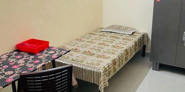 A simple single seater room with bed, chair, table, and cupboard.