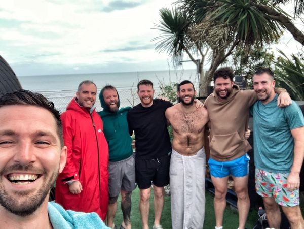 Group of seven men smiling outdoors by the sea, some in swimwear and towels.