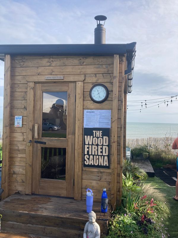 A wooden wood-fired sauna by the seaside with a clock and garden statue.