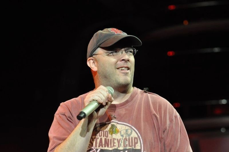 Man speaking into a microphone wearing a cap and glasses.
