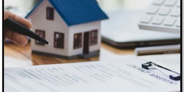 A hand holding a pen near a contract with a miniature house model in the background.